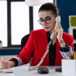 Businesswoman taking notes on clipboard sitting at desk in corporate office, while taking with adviser about financial expertise. Busy manager having a conversation about deadline.