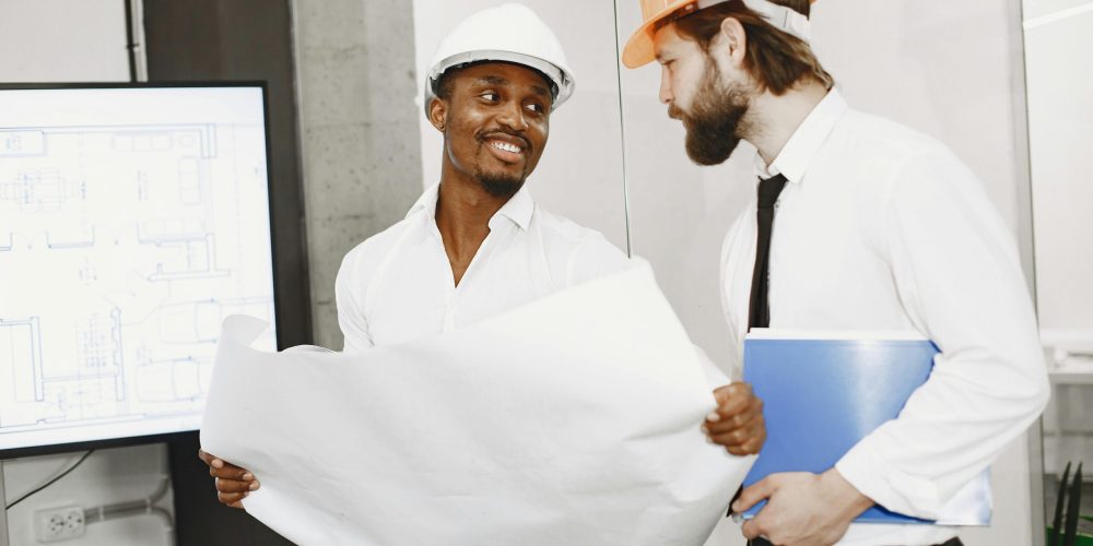 Two architects in hard hats discuss blueprints in a modern office setting, highlighting teamwork and planning.